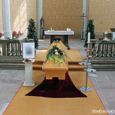 Holz-Sarg mit Blumenarrangement auf rotem Teppich in einer Kirche, umgeben von Bänken und Kerzenständern.
