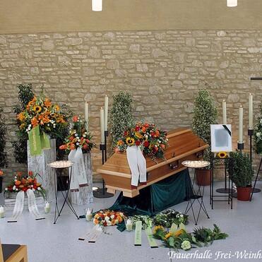 Holzkorb in einer Trauerhalle, umgeben von Blumenarrangements, Kerzen und einem Kreuz, mit einer ruhigen Atmosphäre.
